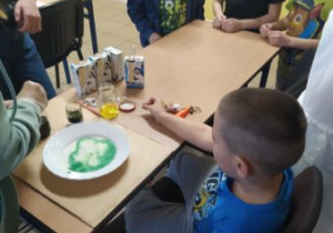 Zainteresowani chłopcy stoja przy stoliku. Na stoliku ustawiono kartoniki z mlekiem , barwniki i talerz z mlekiem. Po mleku pływają zielone esy -floresy.